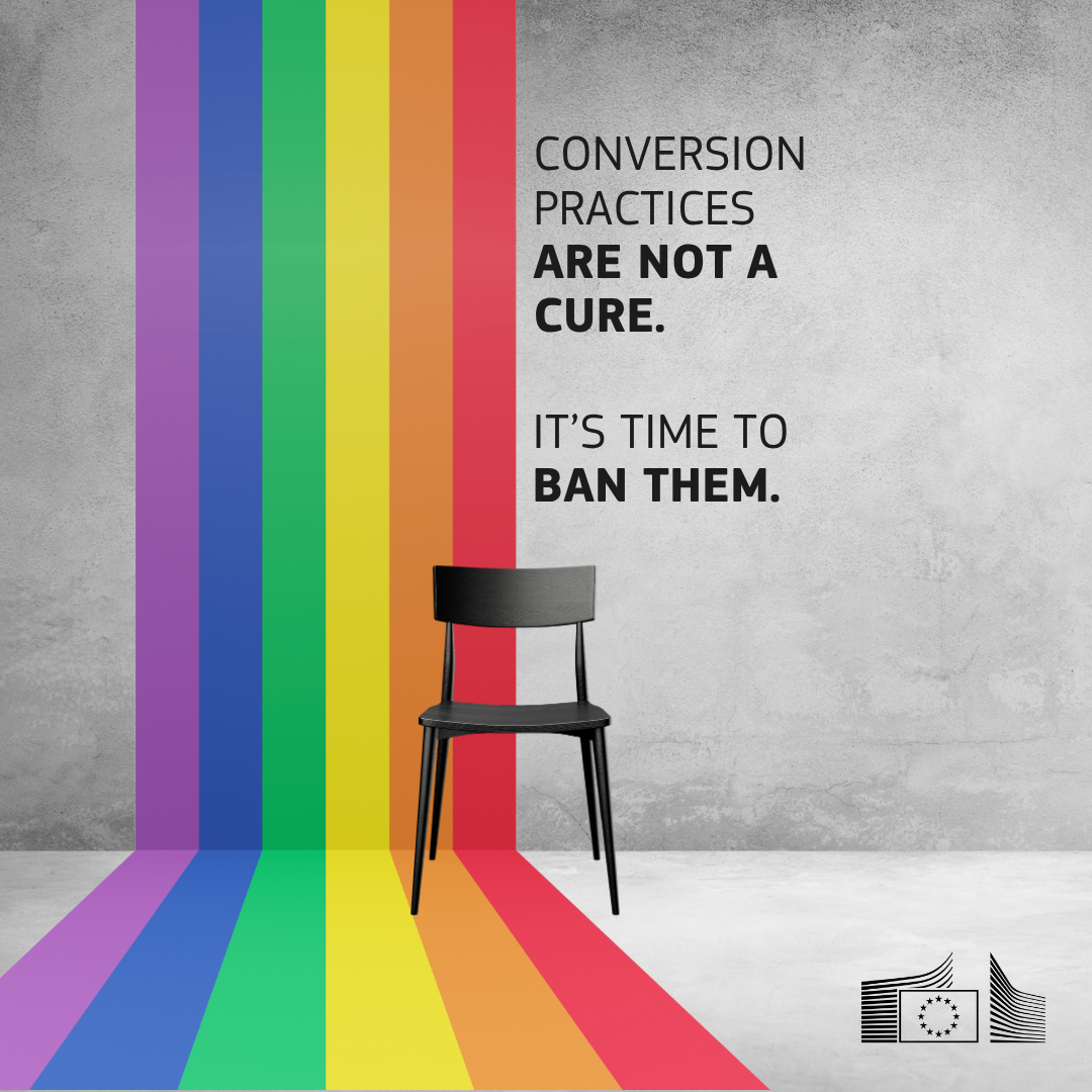 Grey painted room with a rainbow flag running from the top of the wall through the ground. A black chair stands between the flag and the grey wall. The text reads: ‘Conversion practices are not a cure. It’s time to ban them’.